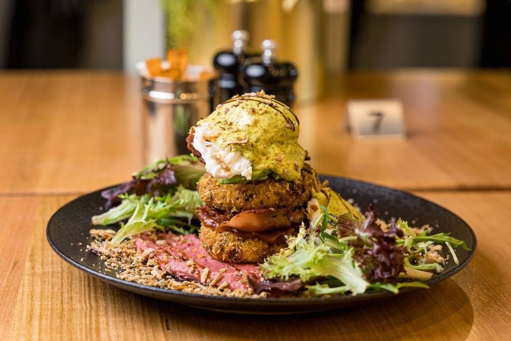 Stacked crumbed breakfast hash with poached egg and herb sauce on a dark plate with sliced beef