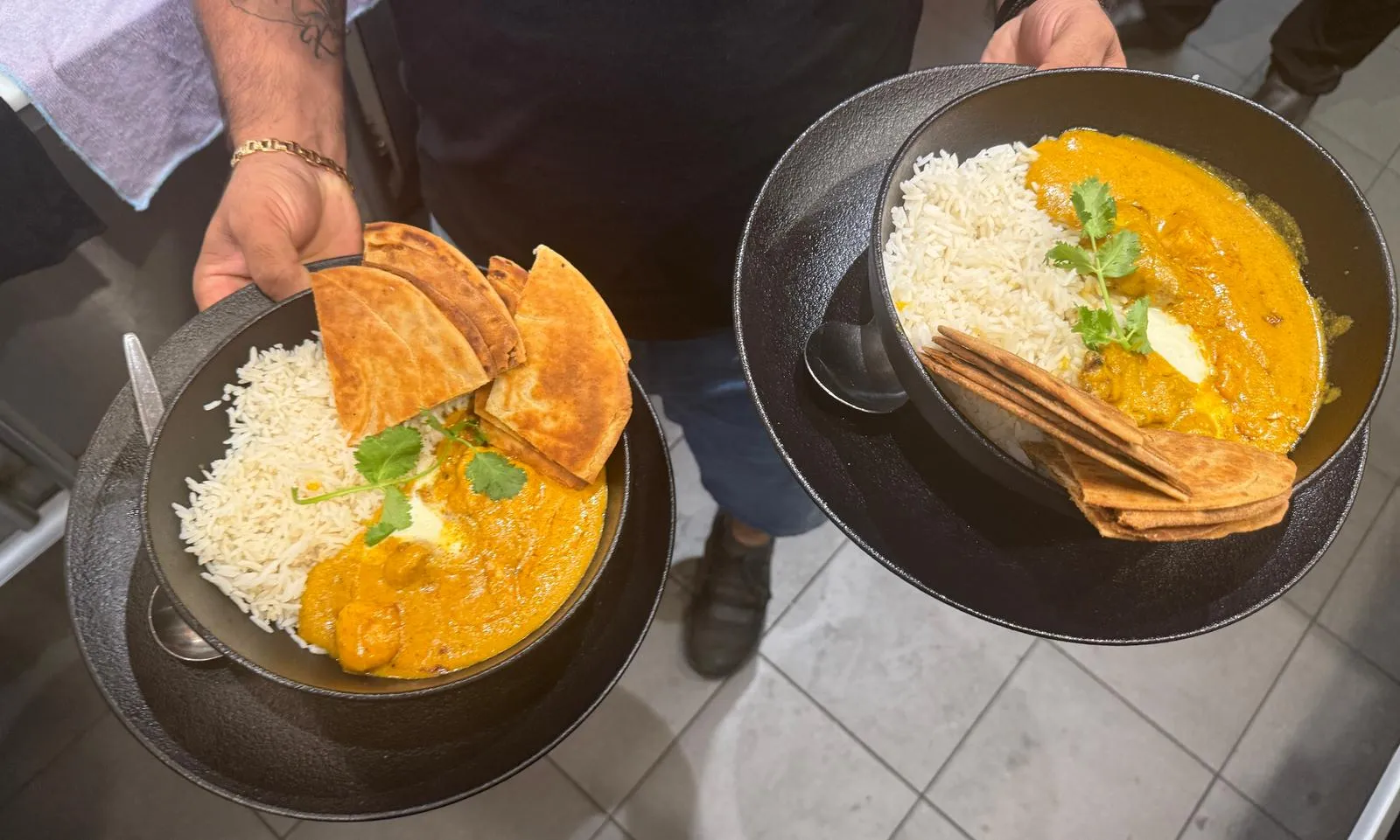 Staff hands holding two black bowls of yellow curry with rice, coriander and flatbread