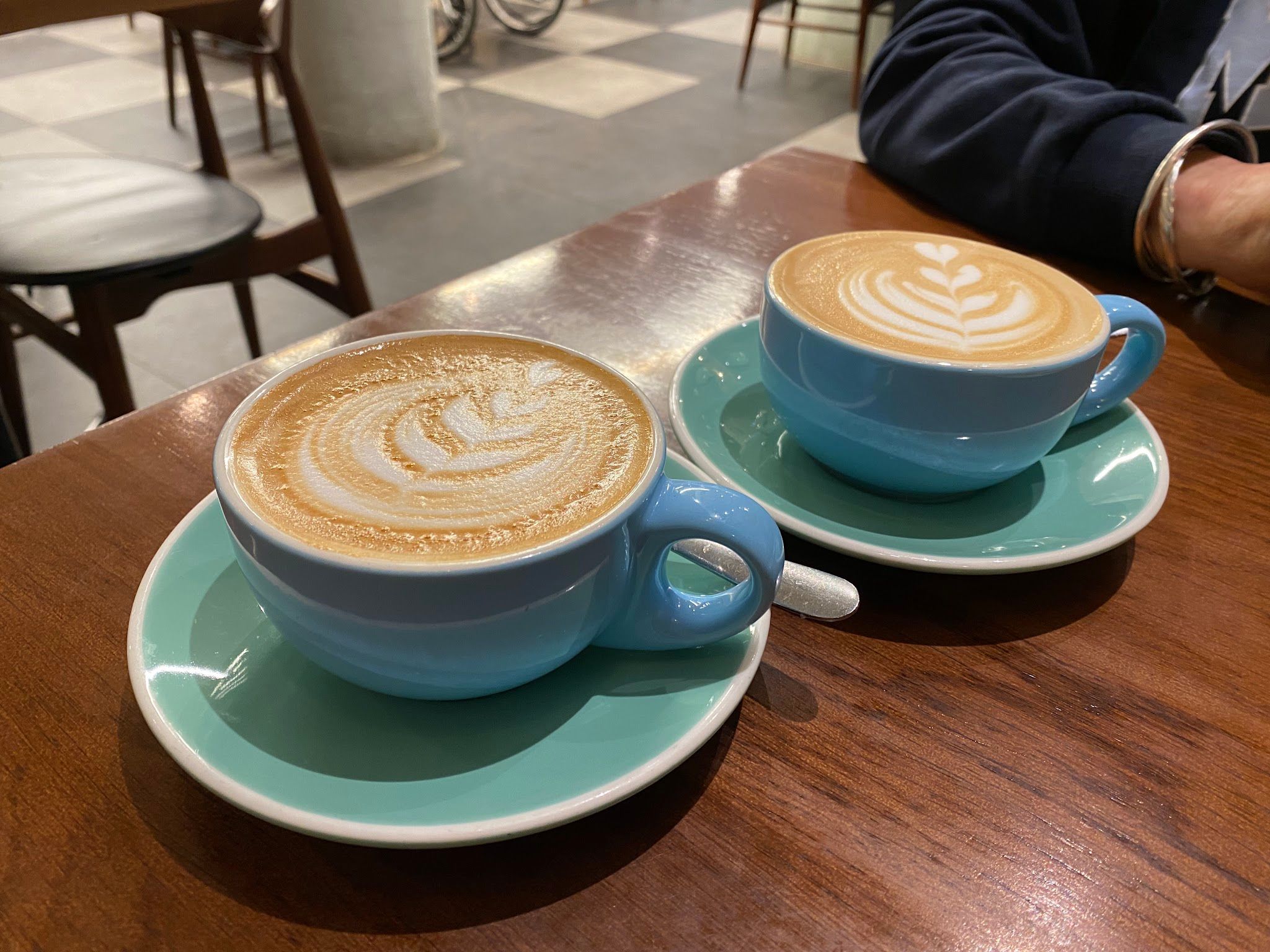 Two blue ceramic cups of latte art on saucers on a café table with checkerboard floor in background