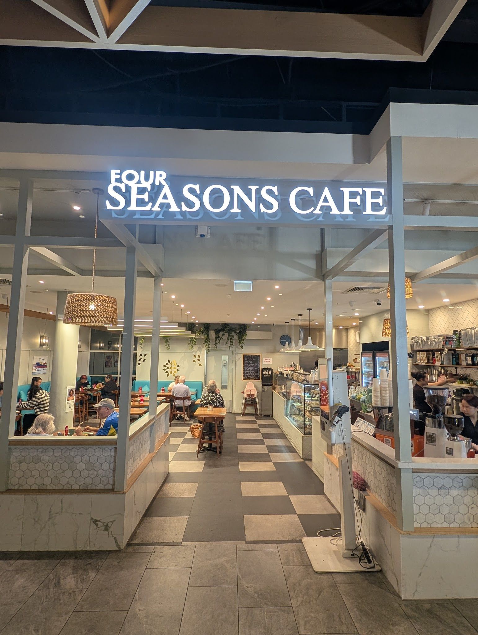 Four Seasons Cafe lit shopfront at evening with glass frontage showing customers inside and checkerboard floor