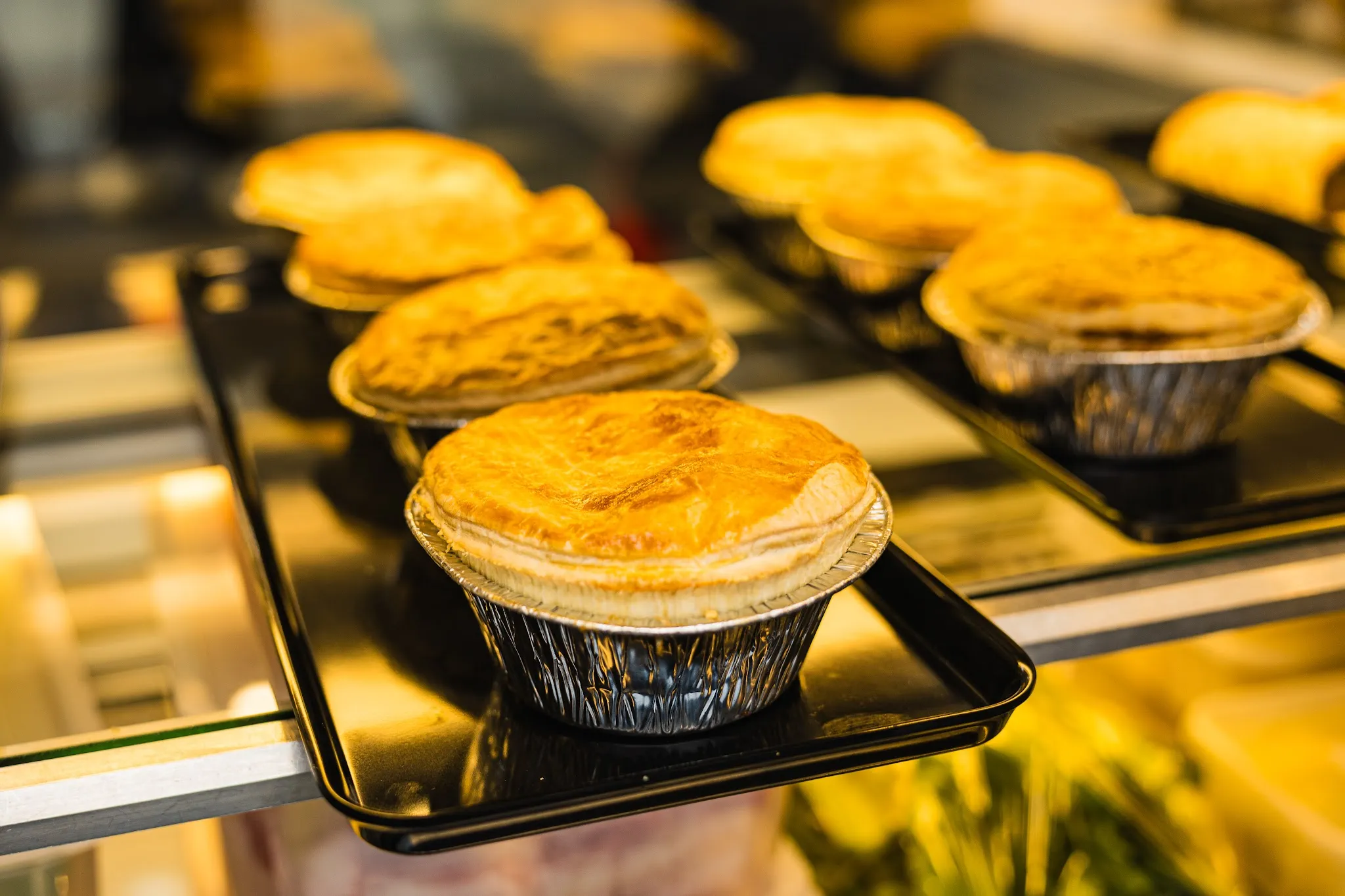Golden-brown meat pies in foil tins on a black tray under warm display lighting