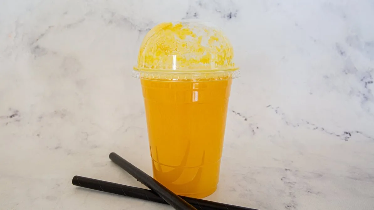 Iced mango drink in a clear dome-lid cup with crushed ice and black straws on marble